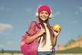 Happy girl in casual fashion style listen to music in modern headphones holding apple fruit for travel sunny outdoors
