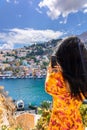 Happy girl enjoys view of coast of Symi Islands, Greece. Woman in red taking photo on phone of colorful houses, blue sea Royalty Free Stock Photo