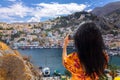 Happy girl enjoys view of coast of Symi Islands, Greece. Woman in red taking photo on phone of colorful houses, blue sea Royalty Free Stock Photo