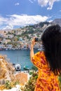 Happy girl enjoys view of coast of Symi Islands, Greece. Woman in red taking photo on phone of colorful houses, blue sea Royalty Free Stock Photo