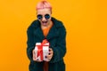 Happy girl with pink hair with a nose and tongue piercing dressed in a fur coat and glasses holds a gift on a yellow Royalty Free Stock Photo