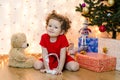 Happy girl in red festive clothes with hat under the Christmas tree waits 12 hours and presents from Santa Claus. Royalty Free Stock Photo