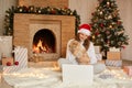 Happy girl in red hat having video call on laptop and sitting with cute dog at christmas tree with lights and presents in festive Royalty Free Stock Photo