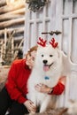 Happy girl in red new year sweater sits on the stairs and hugging cute white Samoyed dog with small red deer horns Royalty Free Stock Photo