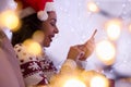 Happy girl with Santa Claus hat and Christmas sweater is shopping online using smartphone at home. blurred lights. Royalty Free Stock Photo