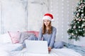 Happy girl in santa hat shopping online on laptop and sitting on bed next Royalty Free Stock Photo