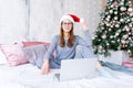 Happy girl in santa hat shopping online on laptop and sitting on bed next Royalty Free Stock Photo