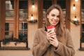 Happy girl stands in the street with a cup of coffee in her hands, looks at the camera, smiles, poses on the camera.Smiling girl