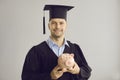 Happy graduate in cap and gown holding piggy bank with money for postgraduate education