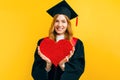 A happy graduate in a master`s dress holds a paper red heart on a yellow background Royalty Free Stock Photo