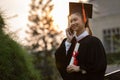 Happy graduated young woman in cap and gown talking with parent in mobile phone and looking certificated in hand so proud