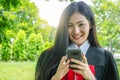 Happy graduated young woman in gown talking with parent in mobile phone and looking camera with nature background