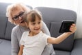 Happy grandmother and granddaughter using phone together, sitting on cozy sofa at home, browsing mobile device apps Royalty Free Stock Photo