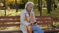 Happy grey-haired beautiful mature woman using smartphone online in autumn park Caucasian female using internet Royalty Free Stock Photo