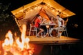 Happy group of friends relaxing in glamping and drinking wine on summer evening near cozy bonfire. Luxury camping tent for outdoor