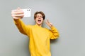 Happy guy in bright casual clothes takes selfie on smartphone on gray wall background and shows peace gesture. Isolated