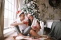 Happy guy and girl in white t-shirts and Santa Claus hats sit with red cups on the floor in front of the window next to Royalty Free Stock Photo