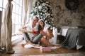 Happy guy and girl in white t-shirts and Santa Claus hats sit with red cups on the floor in front of the window next to Royalty Free Stock Photo