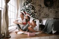 Happy guy and girl in white t-shirts and Santa Claus hats sit with red cups on the floor in front of the window next to Royalty Free Stock Photo