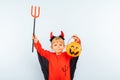Happy Halloween! Cute little boy in devil halloween costume with trident and pumpkin basket jack-o-lantern on light blue Royalty Free Stock Photo