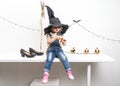 Happy Halloween: A little girl sits on a table in a witch hat with a broom. Royalty Free Stock Photo