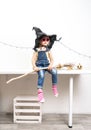 Happy Halloween: A little girl sits on a table in a witch hat with a broom Royalty Free Stock Photo
