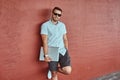 Happy handsome fashionable freelancer in sunglasses dressed in a white shirt and shorts holds a laptop computer standing