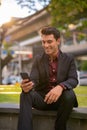 Happy handsome Hispanic businessman using phone and sitting with nature in city Royalty Free Stock Photo