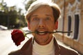 Happy handsome man smiling while holding red rose in mouth standing on street. Valentine`s Day, 8th March, birthday, anniversary Royalty Free Stock Photo