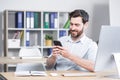 Happy handsome office worker sitting at a computer desk in the office using a mobile phone. Cheerful bearded business man Royalty Free Stock Photo