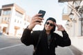 Happy handsome young hipster man in trendy hoodie hooded in stylish sunglasses in a cap with a cute smile makes the selfie