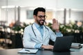 Happy handsome young islamic male doctor with beard in glasses, white coat looks at laptop, waving hand Royalty Free Stock Photo
