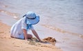 Happy hild girl in big hat and white dress playing alone with wet sand on sandy beach near clear sea lagoon water Royalty Free Stock Photo