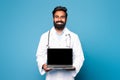 Happy hindu man doctor in white coat with stethoscope showing laptop with blank screen on blue studio background