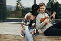 Happy hipster couple relaxing on wooden porch of modern cabin with big windows in mountains. Hipster man playing on ukulele for