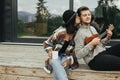 Happy hipster couple relaxing on wooden porch of modern cabin with big windows in mountains. Hipster man playing on ukulele for