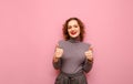 Happy hipster girl with curly red hair stands on a pink background, looks into the camera and shows thumbs up with a smile on her Royalty Free Stock Photo