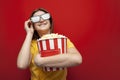 Happy hipster girl with popcorn puts on 3D glasses and smiles on a red colored background Royalty Free Stock Photo