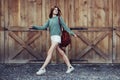 Happy hipster girl walking near barn wooden wall wearing shorts, sweater and brown leather backpack.