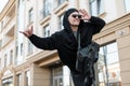 Happy hipster man in sunglasses in stylish black clothes with a leather trendy backpack in a cap waits for a taxi on a street