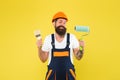 Happy hipster in safety helmet and work uniform hold paint roller and brush for decorating and painting under