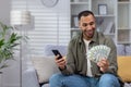 Happy hispanic man holding cash dollars and mobile phone. Sitting at home on the sofa and admiringly looking at the fan