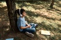 Happy Hispanic teenage boy in headphones, relaxing in the park after school, sitting under tree near scattered textbooks Royalty Free Stock Photo