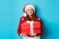 Happy holidays and Christmas concept. Cheerful teen girl with red hair, wearing santa hat and holding xmas presents, Royalty Free Stock Photo