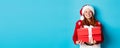 Happy holidays and Christmas concept. Cheerful teen girl with red hair, wearing santa hat and holding xmas presents, Royalty Free Stock Photo