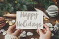 Happy Holidays text on greeting card in hands and christmas ornaments, decorations, confett, branches. Season`s greetings card. Royalty Free Stock Photo