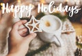 Happy holidays text handwritten on hand holding christmas star gingerbread cookie at cozy  sweater, coffee, fir branches and Royalty Free Stock Photo