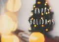 Happy Holidays text sign on blurred christmas tree and golden lights bokeh in festive white room. Winter holidays preparation. Royalty Free Stock Photo