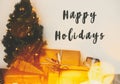 Happy Holidays text sign on christmas tree in golden lights bokeh with festive stylish gifts in white room. Season`s greeting car Royalty Free Stock Photo
