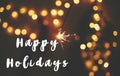 Happy Holidays text sign on glowing sparkler in hand on background of golden christmas tree lights in dark festive room. Fireworks Royalty Free Stock Photo